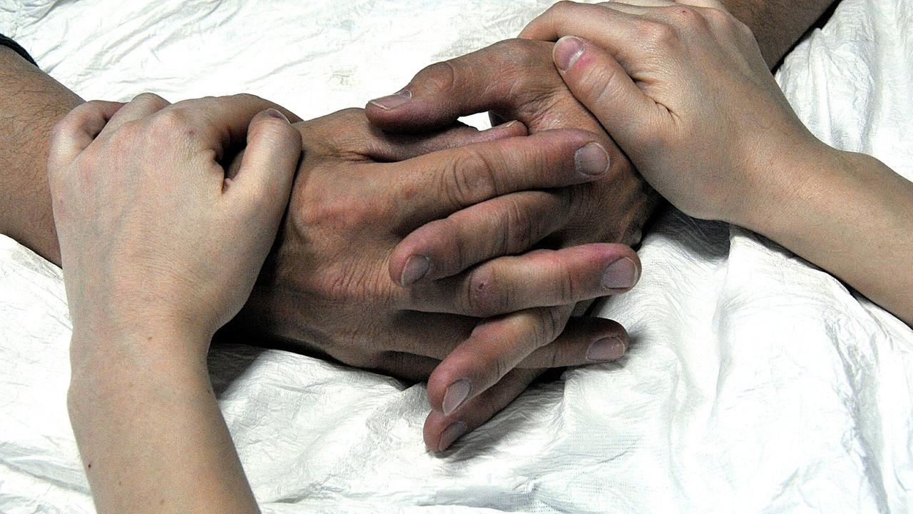 Die Hände eines alten Menschen im Krankenhausbett werden von einer jüngeren Person gehalten.
