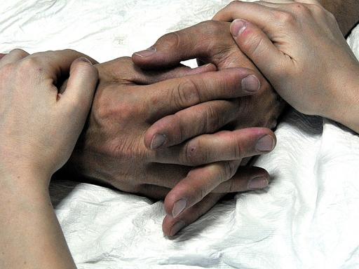 Die Hände eines alten Menschen im Krankenhausbett werden von einer jüngeren Person gehalten.
