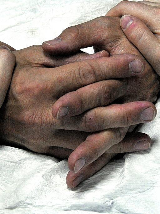 Die Hände eines alten Menschen im Krankenhausbett werden von einer jüngeren Person gehalten.
