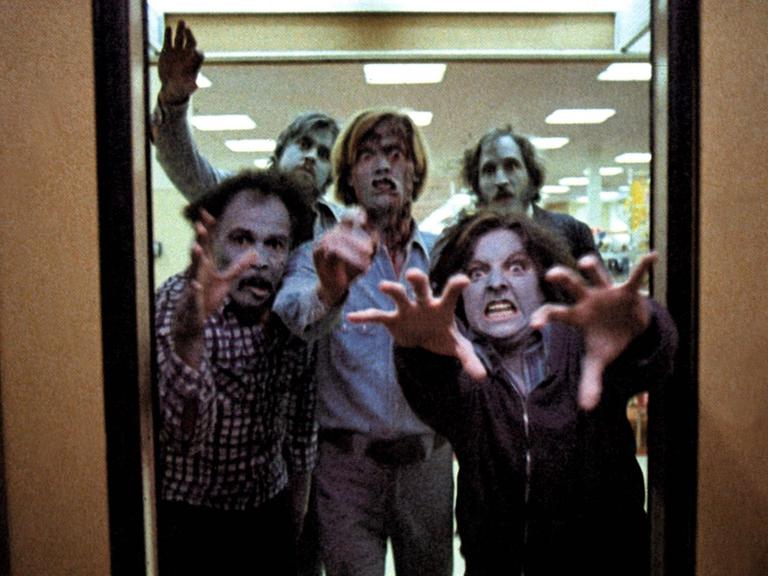 Zombies stürmen durch eine Tür in Richtung Kamera und greifen scheinbar mit den Händen nach dem Betrachter