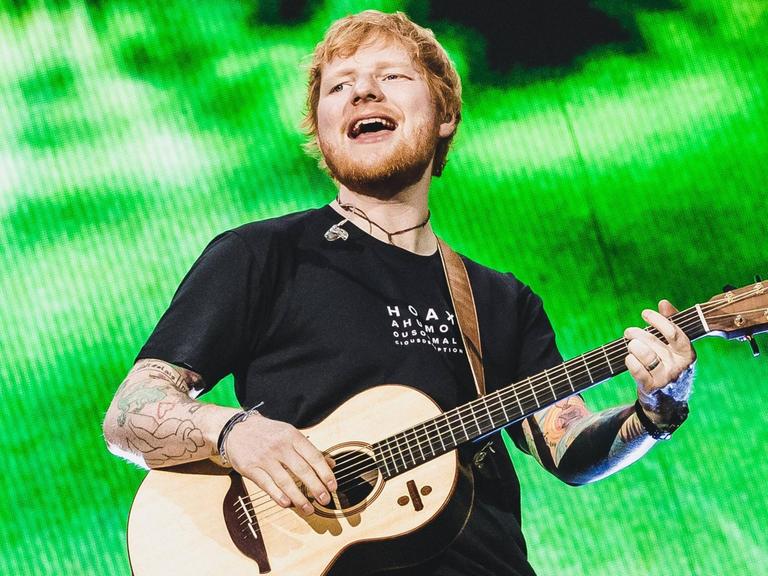 Ed Sheeran steht mit einer Gitarre auf der Bühne, der Hintergrund ist grün.