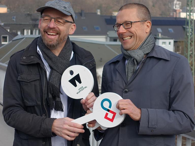 Berthold Schneider (links) und Uwe Schneidewind halten symbolische weiße Schlüssel aus Holz in den Händen, auf denen das Symbol der Oper Wuppertal und des Wuppertal Instituts zu sehen sind.
