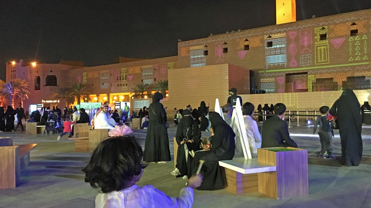 Auf einem Platz in Riad sitzen Männer und Frauen in traditioneller Kleidung auf Bänken.