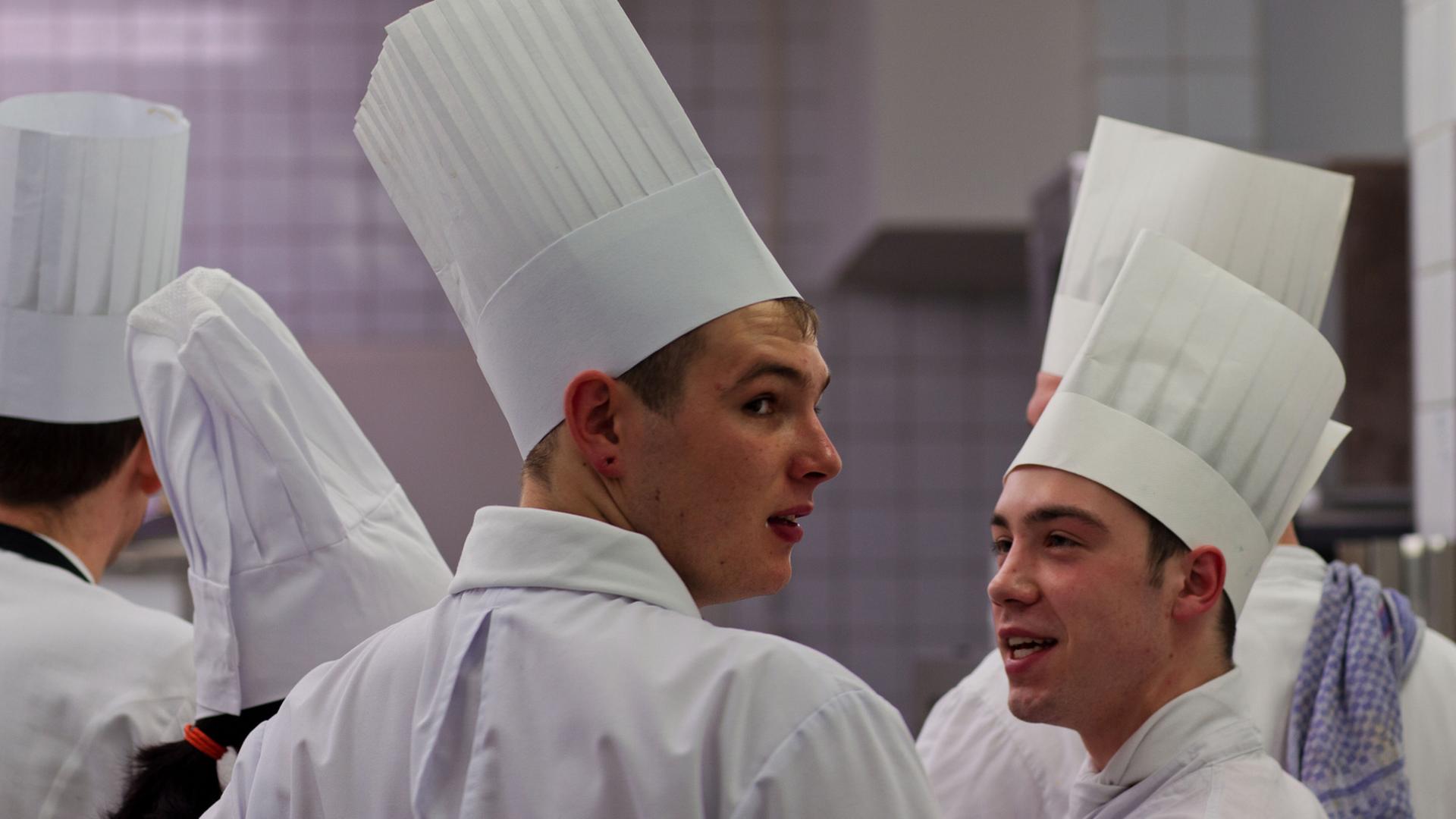 Mehrere Kochlehrlinge mit hohen weißen Kochmützen stehen bei einem Wettbewerb in einer Großküche.