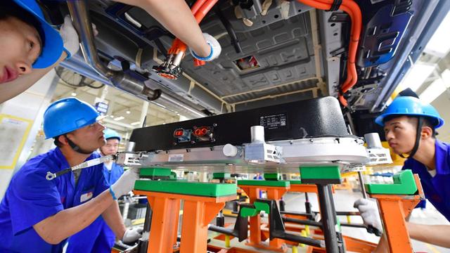 Unteransicht eines Elektroautos, in das eine Batterie installiert wird. Einige chinesische Arbeiter in blauen Fabrikmonturen stehen unter dem Auto, das auf einer Bühne steht. 