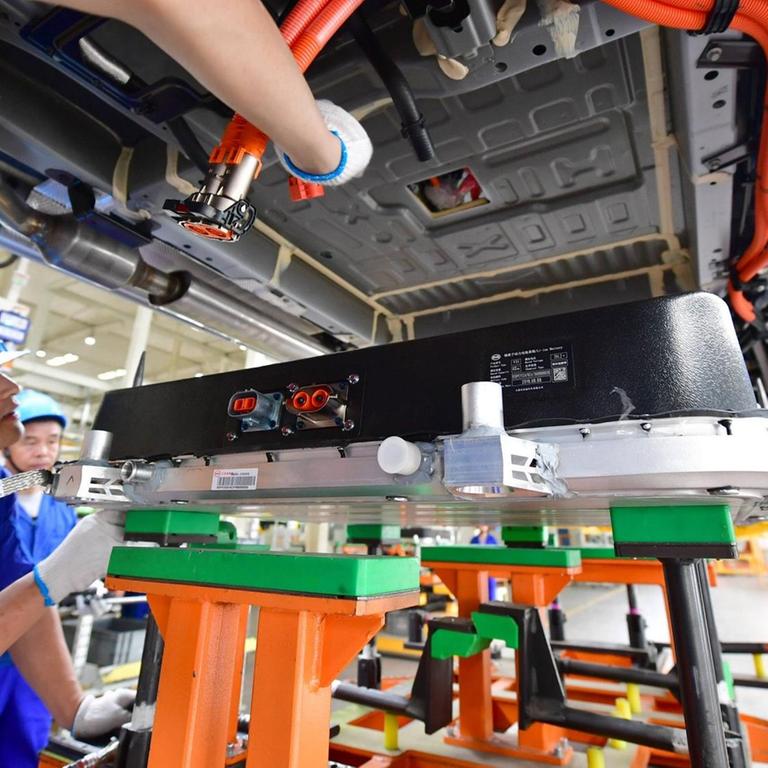Unteransicht eines Elektroautos, in das eine Batterie installiert wird. Einige chinesische Arbeiter in blauen Fabrikmonturen stehen darum und legen Hand an oder beobachten das Geschehen interessiert.
