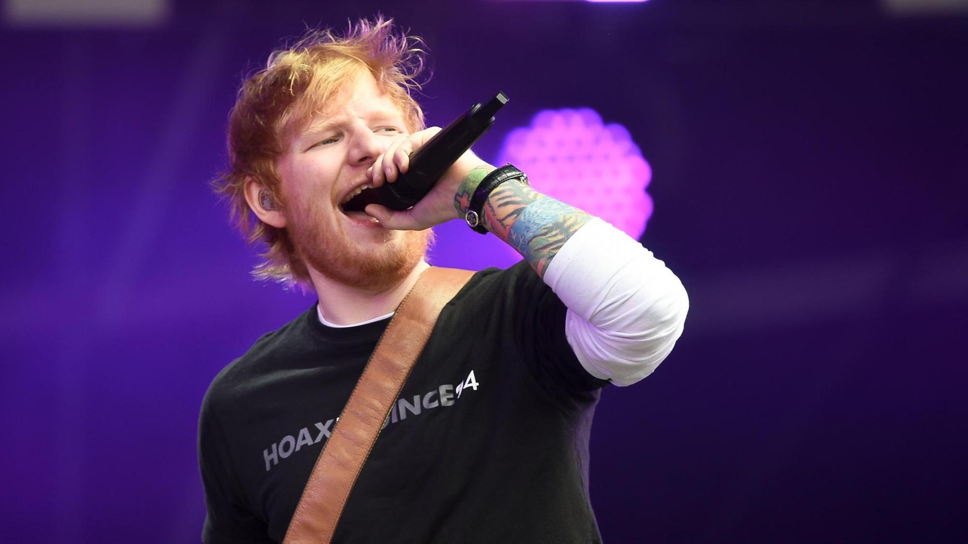 Der Künstler Ed Sheeran hält bei einem Konzert ein Mikrofon in der Hand und singt.