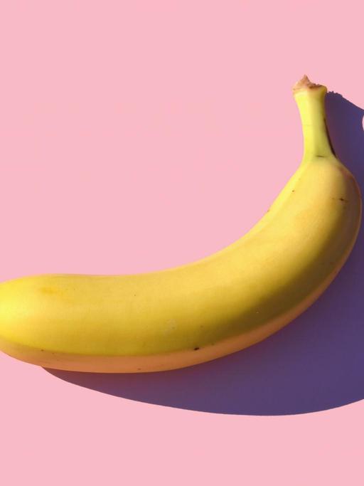 Eine Banane liegt auf einem rosa Untergrund und wirft einen scharfen Schatten darauf.