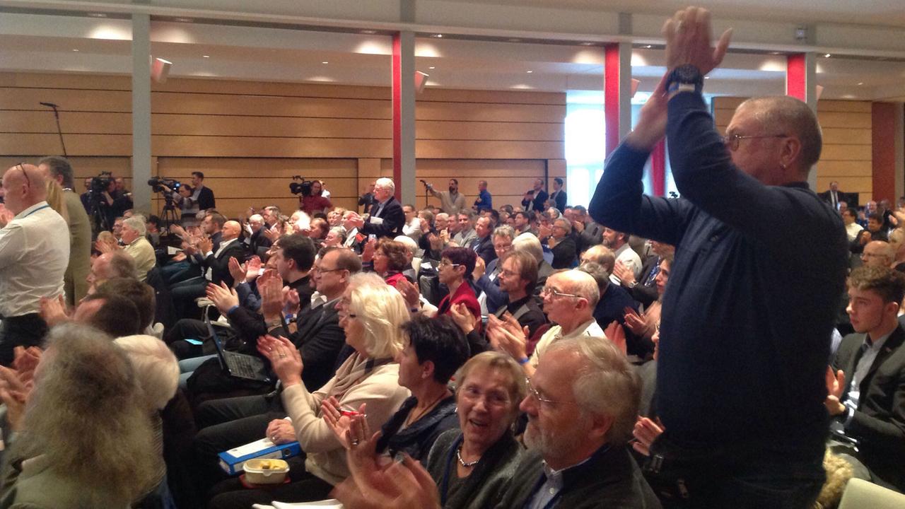 Delegierte der Partei Alternative für Deutschland (AfD) sitzen in Stuhlreihen und applaudieren. Einige klatschen im Stehen.
