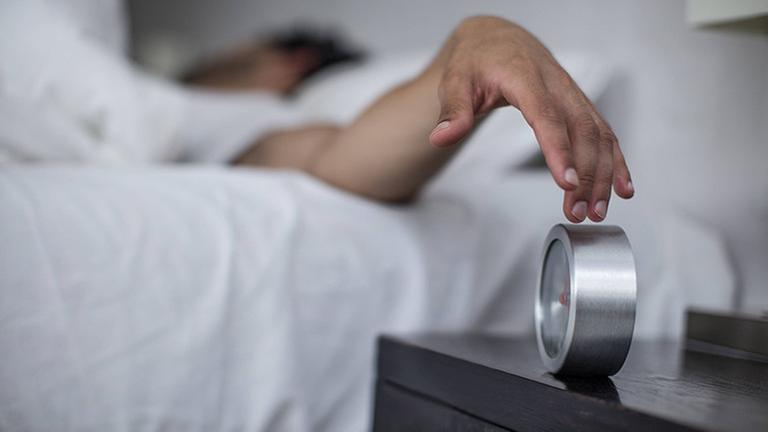 Ein Mann liegt im Bett. Er drückt auf einen Wecker, der neben ihm auf einem Nachttisch steht. Ein Mann liegt im Bett. Er drückt auf einen Wecker, der neben ihm auf einem Nachttisch steht.
