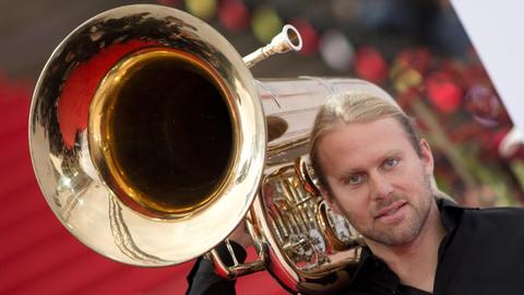 Eine Porträtaufnahme eines blondes Mannes, der eine Tuba schultert, sodass der Betrachter des Bildes in den offenen Trichter blicken kann.
