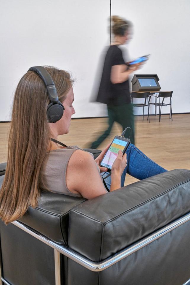 Installationsansicht "Radiophonic Spaces" im Museum Tinguely, Basel. Eine Frau sitzt in einem Sessel, hat einen Kopfhörer auf und schaut auf ihr Smartphone. Im Hintergrund geht eine Frau, ebenfalls einen Kopfhörer auf und schaut auf ihr Smartphon