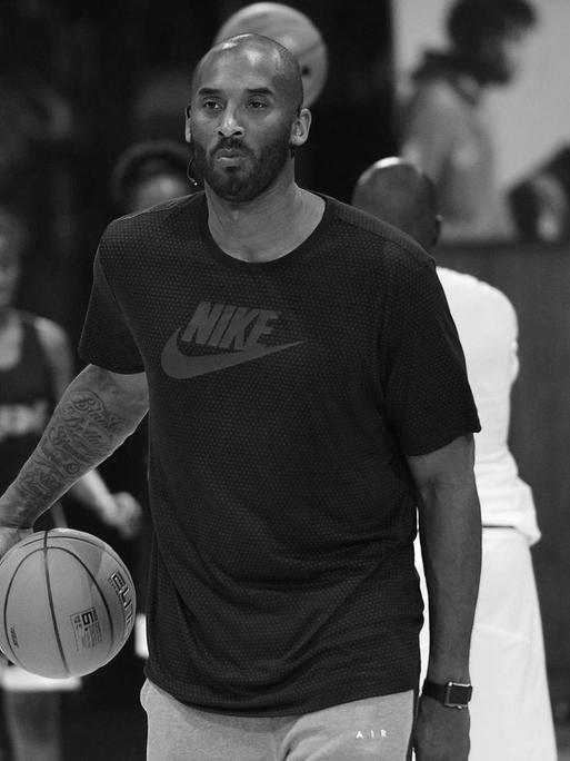 Der Basket-Ball-Spieler Koby Bryant steht auf einem Basket-Ball-Platz und spielt mit einem Ball. Er trägt ein schwarzes T-Shirt und eine graue Sport-Hose.