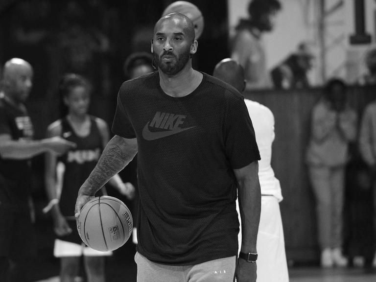 Der Basket-Ball-Spieler Koby Bryant steht auf einem Basket-Ball-Platz und spielt mit einem Ball. Er trägt ein schwarzes T-Shirt und eine graue Sport-Hose.