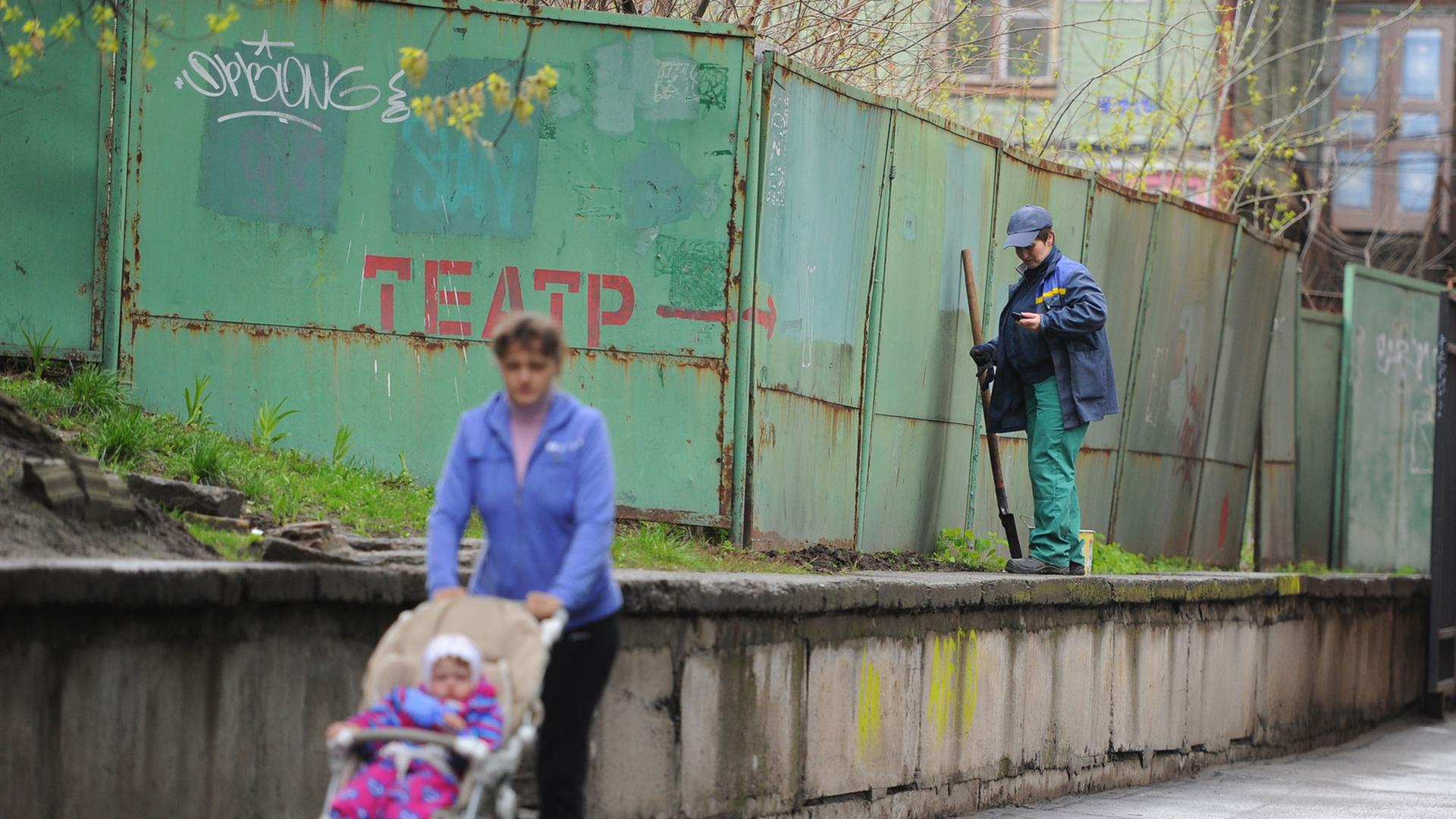 Straßenszene in Kiew - eine Frau geht mit Kinderwagen spazieren, ein Mann reinigt Wände