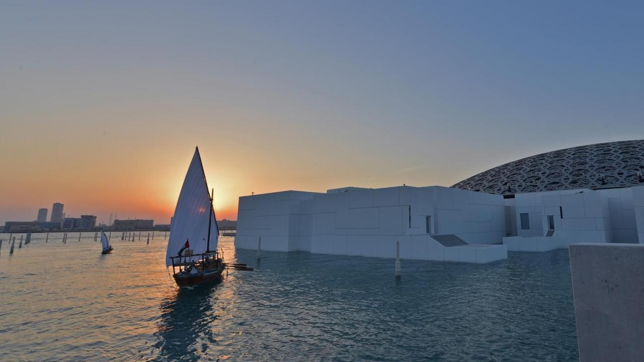 Der Louvre, vom Wasser umspielt, bei Sonnenuntergang mit einem Segelschiff daneben.