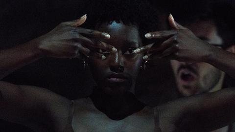 Eine Schwarze Frau hält ihre Hände vors Gesicht und blickt durch die gestreckten Finger in die Kamera. Der Hintergrund und die Umgebung ist sehr dunkel.
