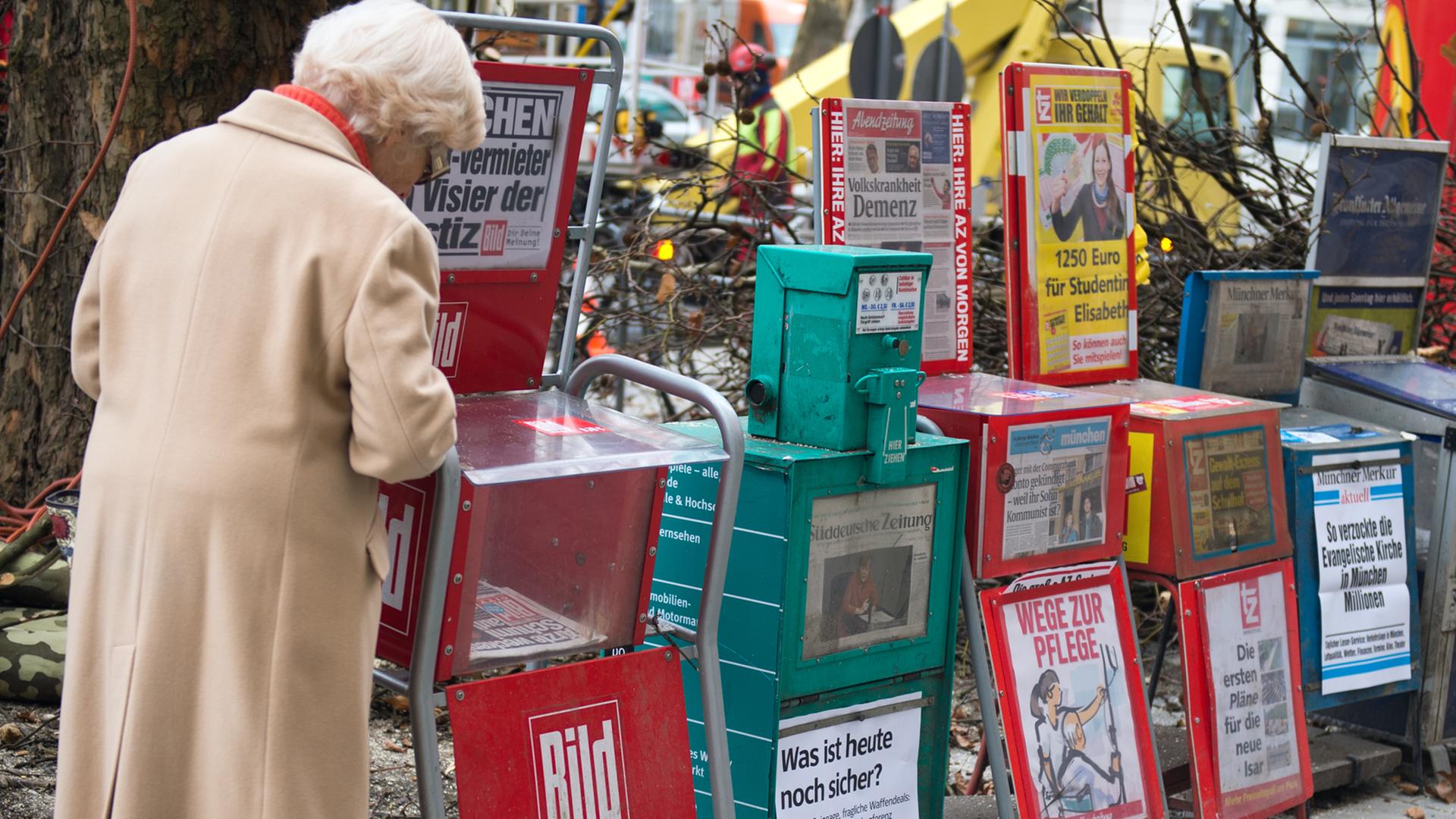 Sechs Zeitungsständer stehen in München am Gehwegrand.