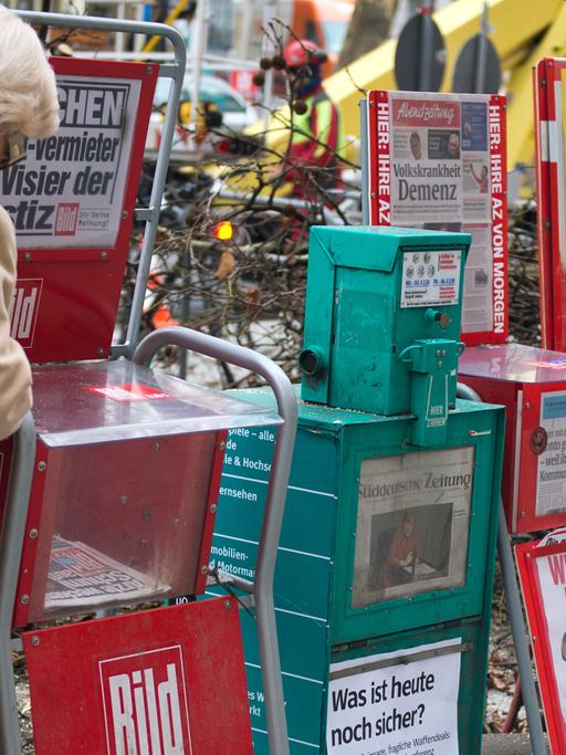 Sechs Zeitungsständer stehen in München am Gehwegrand.