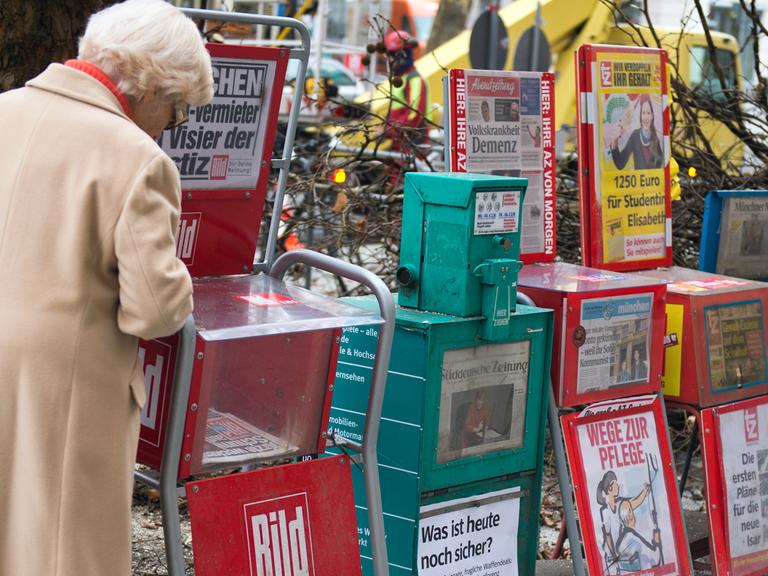 Sechs Zeitungsständer stehen in München am Gehwegrand.