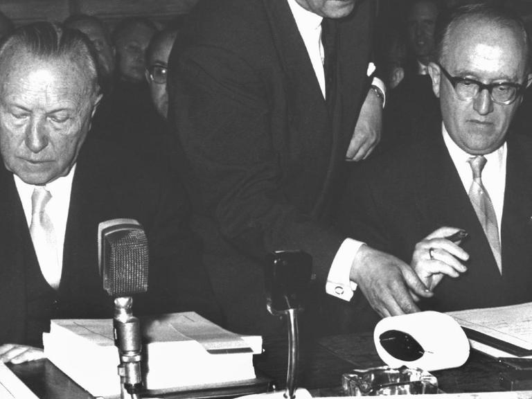Das historische Schwarz-Weiß-Bild zeigt Adenauer und Hallstein nebeneinander an einem Tisch sitzend vor den Verträgen.
