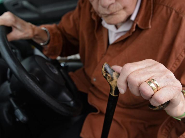 Eine 84-jährige Frau sitzt am Steuer ihres Kleinwagens. In der linken Hand hält sie ihren Gehstock, den sie zum Aussteigen benötigt.