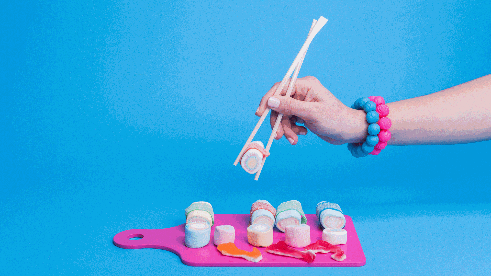 Marshmallows in Suhi-Form geschnitten, liegen auf einem rosafarbenen Brett. Eine Hand hält mit Stäbchen einen Marshmallow.