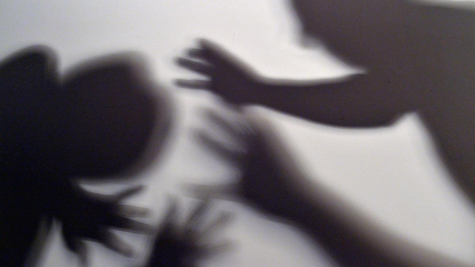 Schatten von Händen und zwei Menschen am Bildrand symbolisieren, wie eine Frau versucht, sich vor der Gewalt eines Mannes zu schützen.