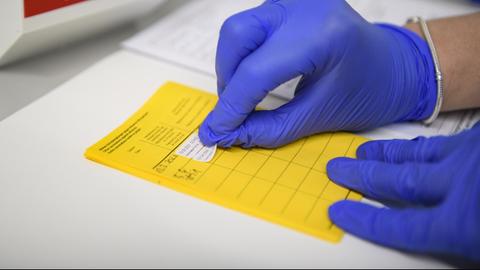 Eine Hand mit Schutzhandschuh klebt einen Aufkleber in einen gelben Impfpass