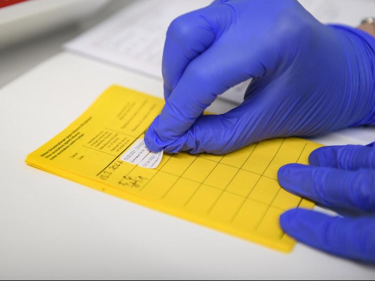 Eine Hand mit Schutzhandschuh klebt einen Aufkleber in einen gelben Impfpass