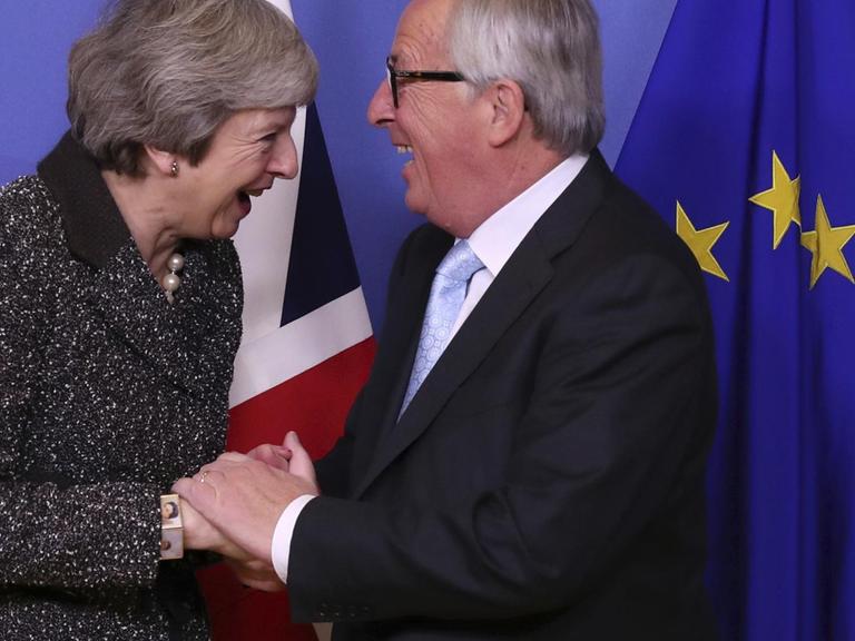 Theresa May und Jean-Claude Juncker halten ihre Hände gegenseitig. Sie lachen herzlich.