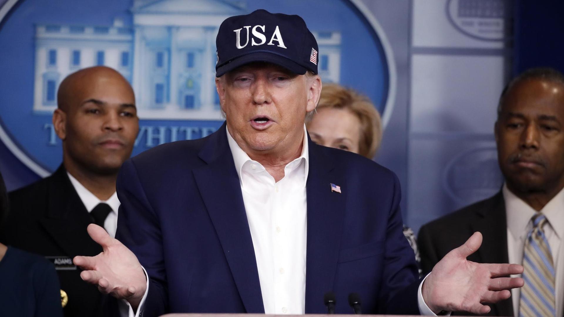 Trump steht in blauer USA-Baseballmütze am Rednerpult im Presseraum des Weißen Hauses. Hinter ihm stehen zwei Männer und eine Frau.