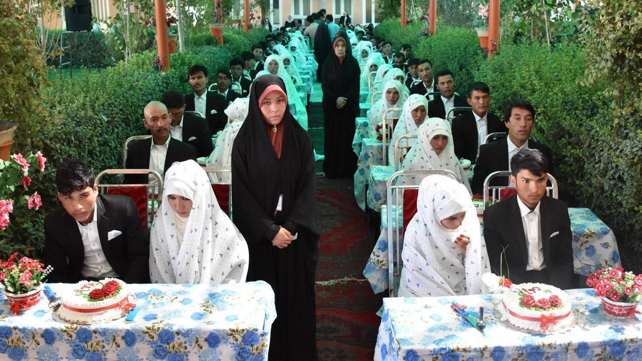 Gruppenhochzeit in Masar-e-Sharif in Afghanistan, aufgenommen im August 2018: Brautpaare - alle gleich gekleidet, die Männer in schwarzen Anzügen, die Frauen weiß verschleiert - sitzen an identisch dekorierten Tischen mit Hochzeitstorten hintereinander, und gucken ernst Richtung Kamera