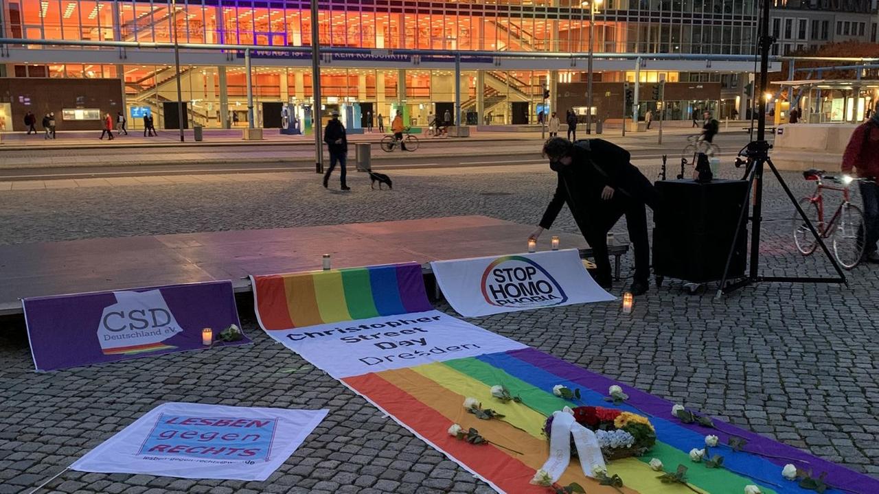 Eine Regenbogenfahne, ein Kranz und weiße Blumen erinnern auf dem Altmarkt vor dem Kulturpalast in Dresden an die Opfer einer Messerattacke