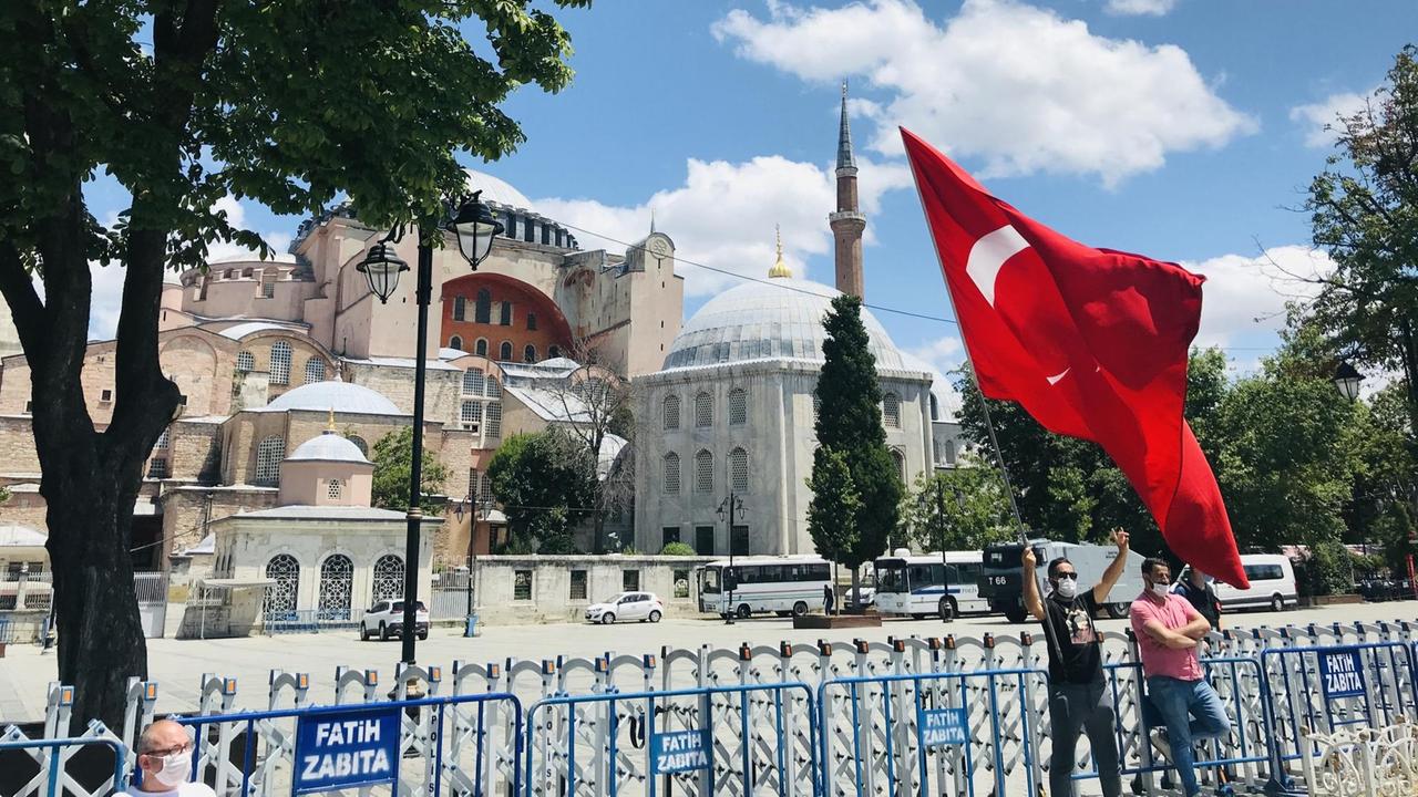Ein Mann mit Mundschutz steht vor den Absperrungen vor der Hagia Sophia und schwenkt die rote Fahne der Türkei.