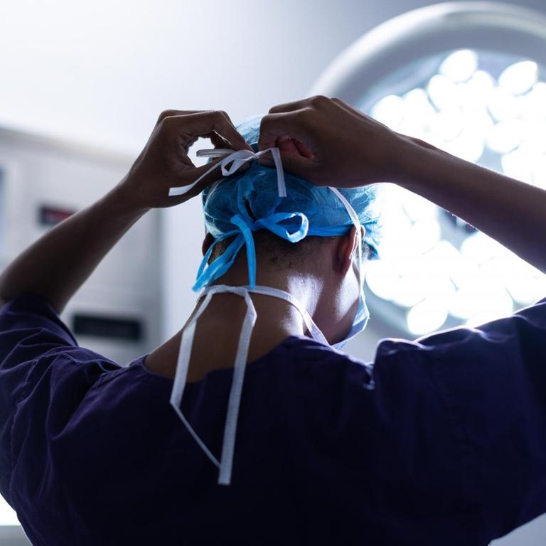 Eine Chirurgin ist im Operationssaal eines Krankenhauses von hinten zu sehen, während sie sich eine OP-Maske zubindet. 