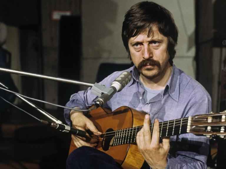 Wolf Biermann sitzt mit Gitarre vor einem Mikrofon.