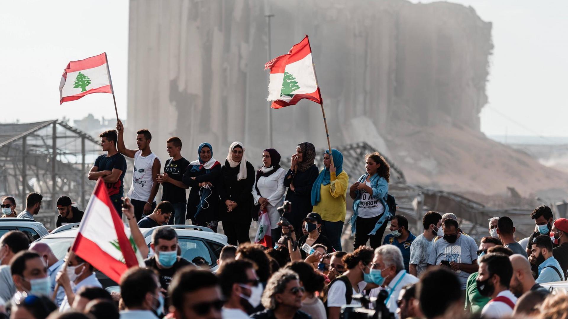 Menschen haben sich vor dem zerstörten Hafen versammelt, sie schwenken Nationalflaggen