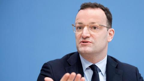 Bundesgesundheitsminister Jens Spahn bei einer Pressekonferenz: Der Minister steht in schwarzem Anzug, weißem Hemd und dunkler Krawatte vor einem mittelblauen Hintergrund und hält beide Handflächen offen nach vorn.