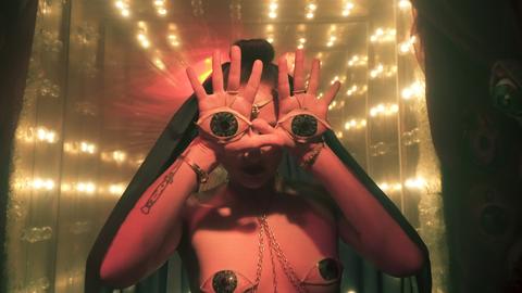 Szenenbild aus der Ausstellung "Overmorrow" im Berliner Club "Wilde Renate": Brustbild einer Frau, die die Hände mit gespreizten Fingern vor ihr Gesicht hält, auf den Handflächen sieht man Augen, im Hintergrund strahlenförmige Lichter.