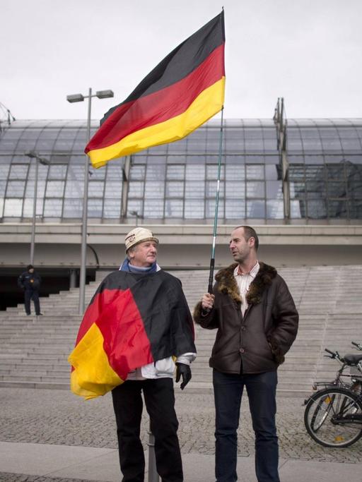 Zwei Männer stehen vor einer grauen Steintreppe, einer hält eine Deutschland-Fahne in der Hand, der andere hat eine Deutschland-Fahne um den Oberkörper gewickelt.