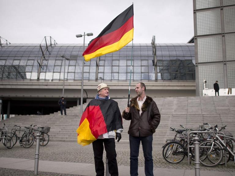 Zwei Männer stehen vor einer grauen Steintreppe, einer hält eine Deutschland-Fahne in der Hand, der andere hat eine Deutschland-Fahne um den Oberkörper gewickelt.