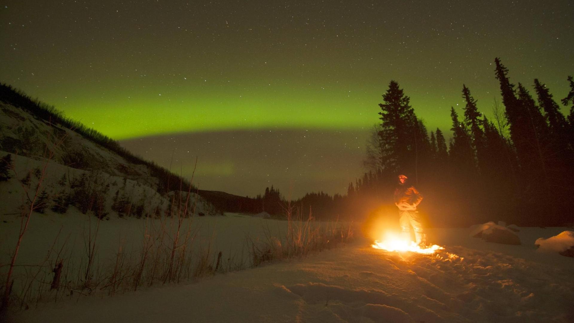 Ein Manns steht außerhalb von Fairbanks/Alaska am Lagerfeuer und beobachtet das Polarlicht.