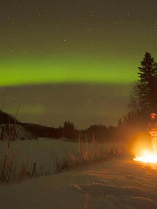 Ein Manns steht außerhalb von Fairbanks/Alaska am Lagerfeuer und beobachtet das Polarlicht.