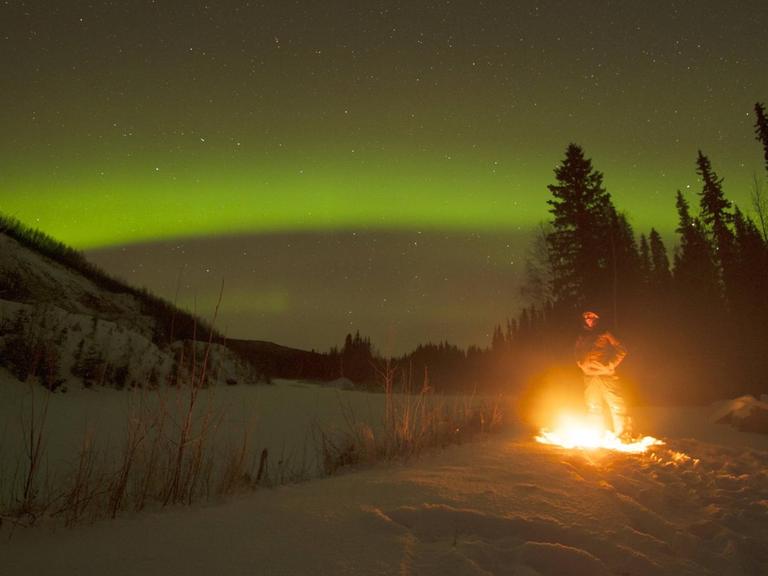 Ein Manns steht außerhalb von Fairbanks/Alaska am Lagerfeuer und beobachtet das Polarlicht.
