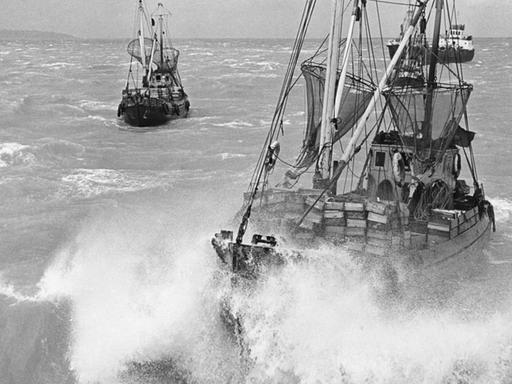 Zwei große Fischerboote mit hohen Masten und eingerollten Segeln befinden sich inmitten tosender Wellen.