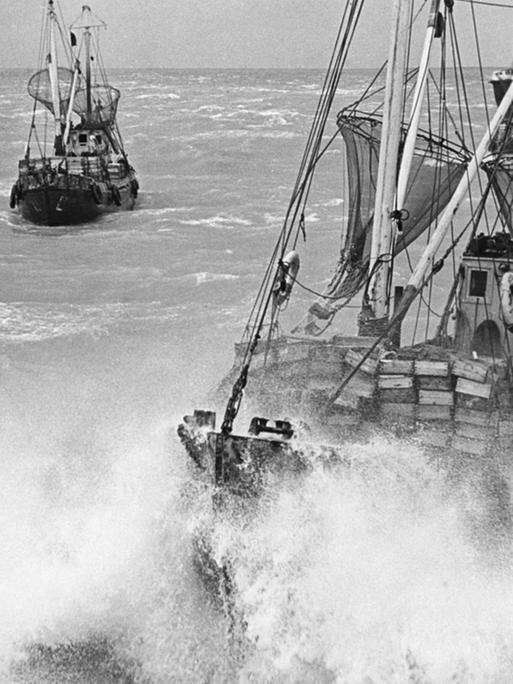 Zwei große Fischerboote mit hohen Masten und eingerollten Segeln befinden sich inmitten tosender Wellen.