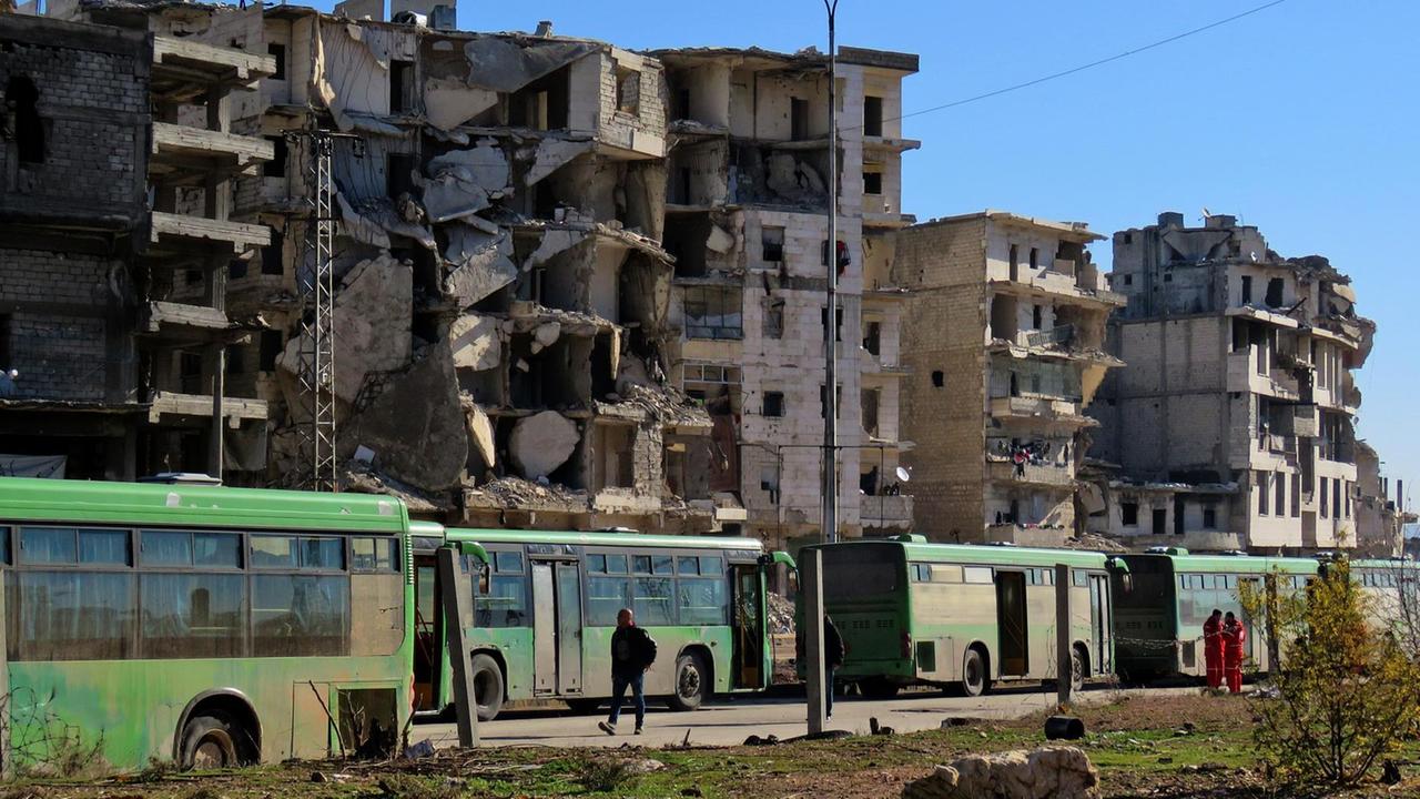 Mehrere grüne Busse fahren in einer langen Reihe hintereinander. Auf beiden Seiten der Straße sind zerstörte Gebäude. Mehrere grüne Busse fahren in einer langen Reihe hintereinander. Auf beiden Seiten der Straße sind zerstörte Gebäude.