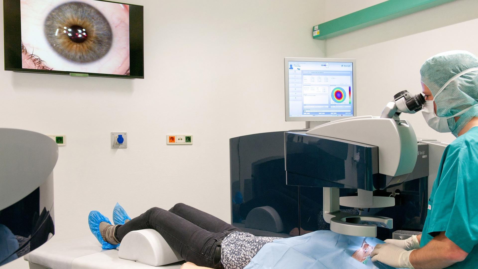 In einer Augenklinik wird eine Patientin auf dem Stuhl liegend behandelt. Die OP wird auf einen Bildschirm an der Wand übertragen.