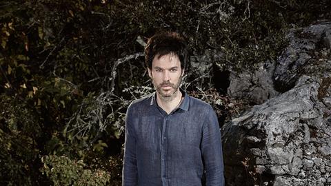 Ein Mann in blauem Jeanshemd, mit schwarzen Haaren und leicht grauem Bart steht bei Dunkelheit vor einem Felsen, der das Blitzlicht reflektiert.